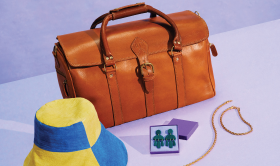 Still life photograph of a brown leather travel bag, yellow and blue paneled bucket hat, a set of dark emerald colored tiered earrings in a purple jewelry box, and a gold braided necklace and bracelet set on a lavender background with a small Etsy logo in black font in the top left corner
