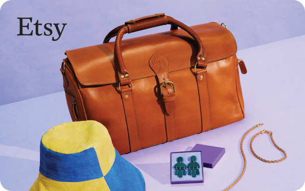 Still life photograph of a brown leather travel bag, yellow and blue paneled bucket hat, a set of dark emerald colored tiered earrings in a purple jewelry box, and a gold braided necklace and bracelet set on a lavender background with a small Etsy logo in black font in the top left corner