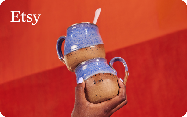 Photograph of a hand holding two stacked handmade ceramic mugs with a natural brown clay base dipped in a light blueish purple glaze on an orange red background with a small Etsy logo in white font in the top left corner