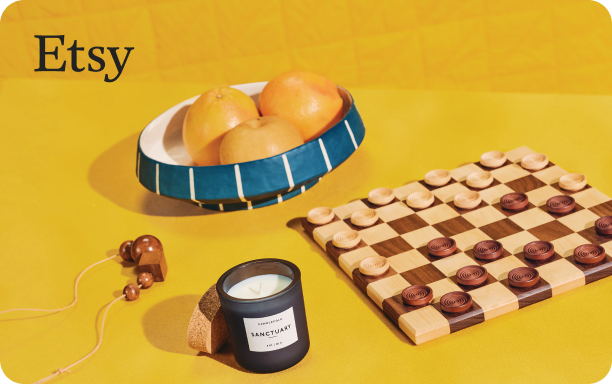 Still life photograph of a checkerboard with wooden pieces on a yellow tabletop. To the left, there’s a candle with the label “L’APOTHECARY” and a cork base. In the background, a blue striped bowl filled with citrus fruit. 